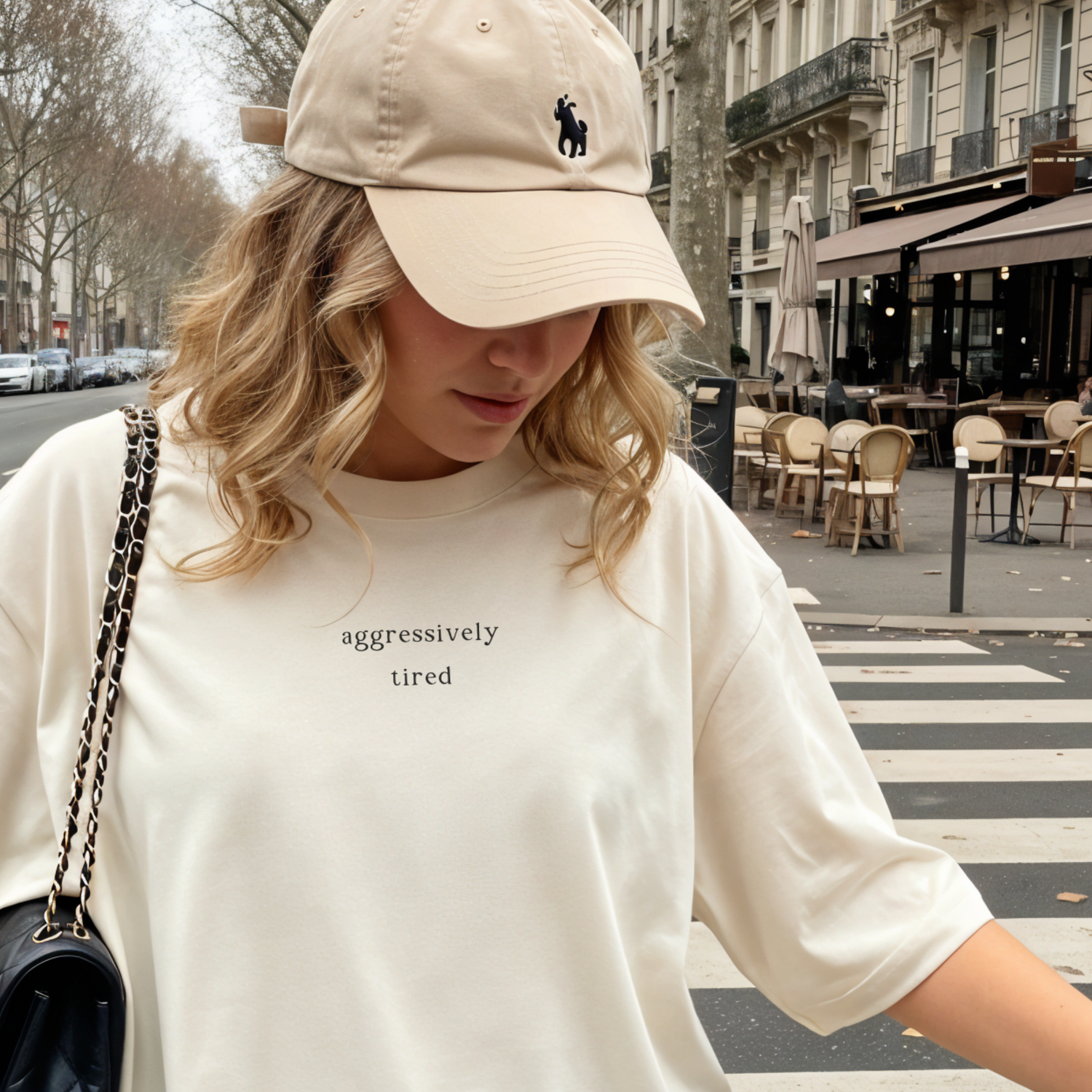 Woman in a cream 'aggressively tired' tshirt and beige baseball cap walking in a city. Aesthetic lifestyle apparel photography for the Barely Sane collection.