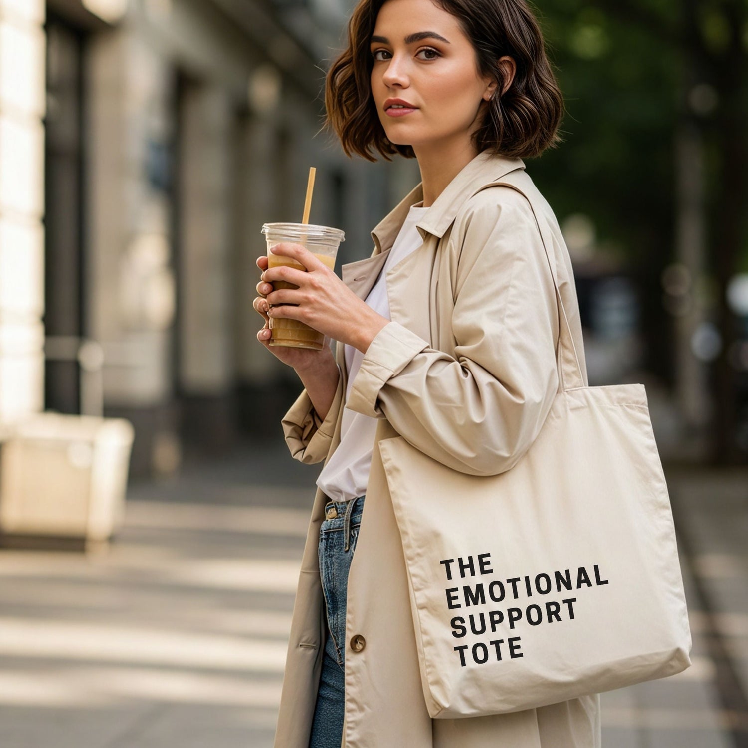 A woman holding an iced coffee and wearing a trench coat, carrying 'The Emotional Support Tote' in natural cotton canvas. Minimalist and aesthetic lifestyle photography for Barely Sane's bag collection.