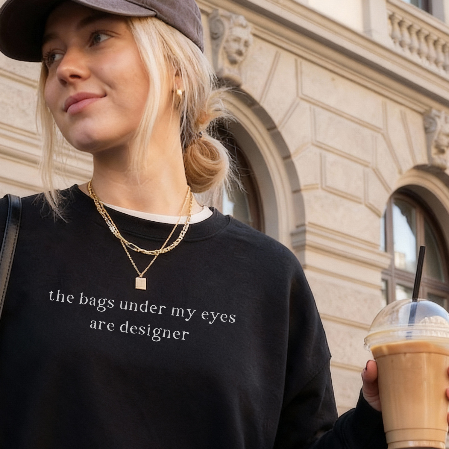 Woman in a black 'The bags under my eyes are designer' sweatshirt and baseball cap holding an iced coffee. Relatable lifestyle apparel by Barely Sane.