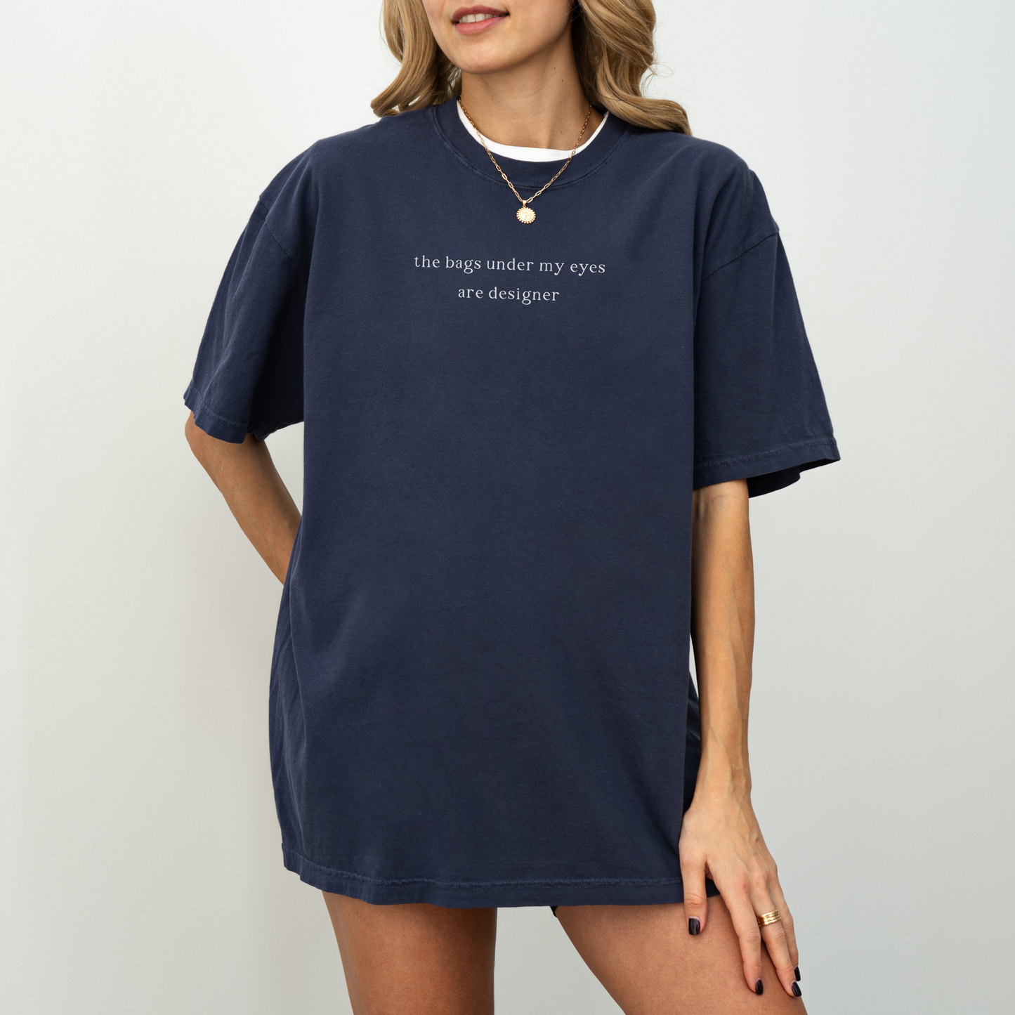 A woman wearing a navy blue 'the bags under my eyes are designer' oversized t-shirt. She is accessorized with a gold necklace and a gold ring. Minimalist studio product photography for the Barely Sane clothing collection.