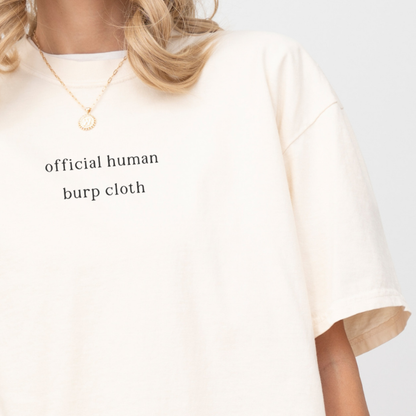 A close-up of a woman wearing an ivory oversized t-shirt with the white text 'official human burp cloth' printed on the chest. She is wearing a gold coin necklace. Minimalist studio product photography for the Barely Sane collection.