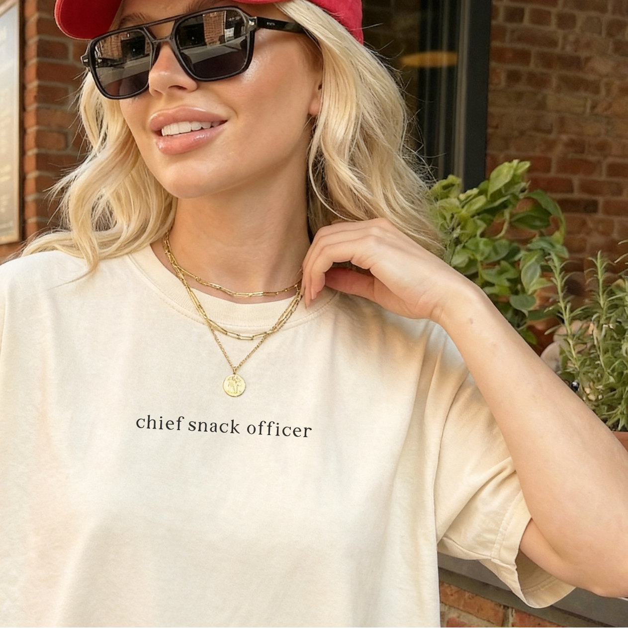 Woman wearing an ivory 'chief snack officer' t-shirt outdoors, paired with sunlasses and a red baseball cap