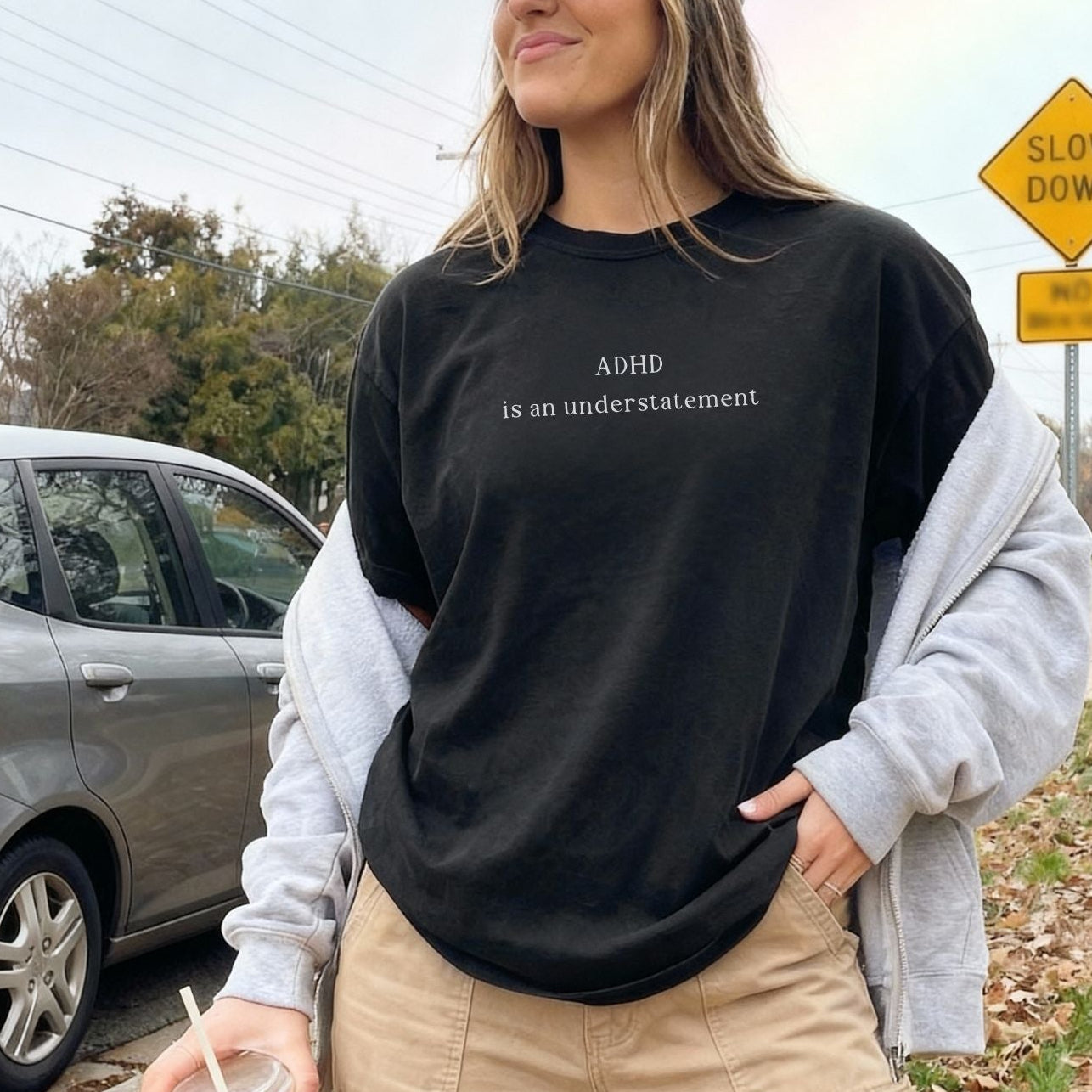 Oversized black t-shirt with white text that reads 'ADHD is an understatement'. Model is wearing a grey zip-up hoodie over the shirt and khaki pants, showcasing the relaxed, aesthetic streetwear look of the graphic tee.