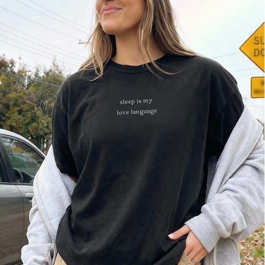 Woman standing on a sidewalk holding a drink, wearing a black tee wthat says 'sleep is my love language'.