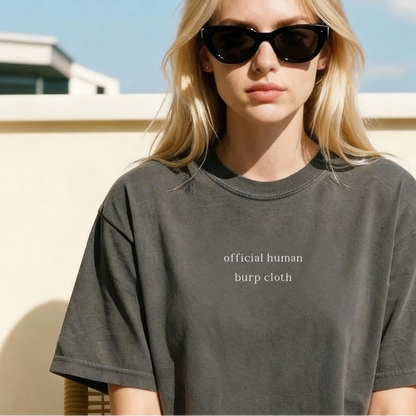 A close-up of a woman wearing sunglasses and a pepper grey oversized t-shirt featuring the white printed text 'official human burp cloth' across the chest. Aesthetic lifestyle photography for the Barely Sane clothing collection.