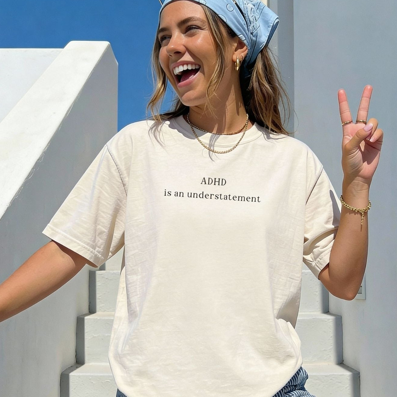 Oversized cream-colored t-shirt with black text that reads 'ADHD is an understatement'. Model is smiling and wearing a light blue bandana, showcasing the casual, aesthetic fit of the graphic tee.