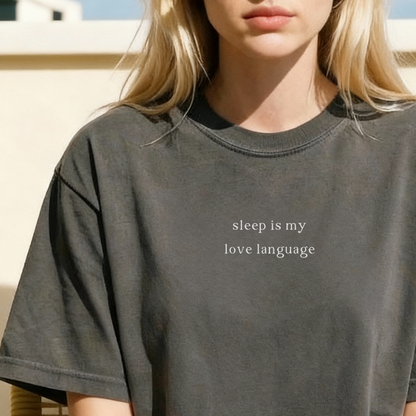 A close-up shot of a woman with blonde hair, wearing a pepper grey oversized t-shirt featuring the white printed text 'sleep is my love language' across the chest.