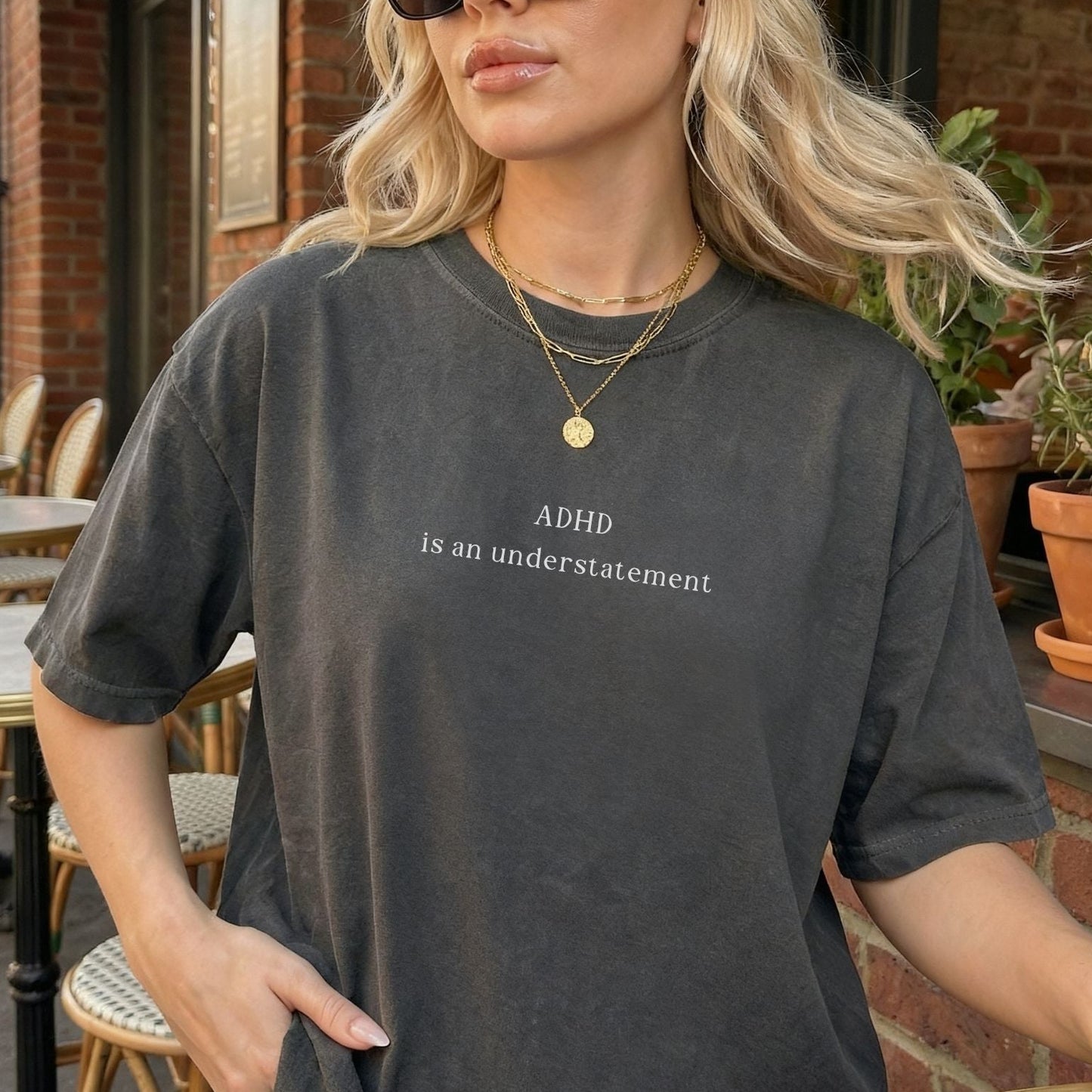 Oversized charcoal grey washed t-shirt with minimalist white text that reads 'ADHD is an understatement'. Blonde model is wearing layered gold necklaces, showcasing the vintage, aesthetic look of the graphic tee at an outdoor cafe.