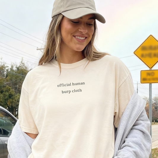 A woman smiling and wearing an ivory 'official human burp cloth' oversized t-shirt, paired with a tan baseball cap and a grey hoodie over her shoulders. Casual, humorous lifestyle photography for the Barely Sane collection.