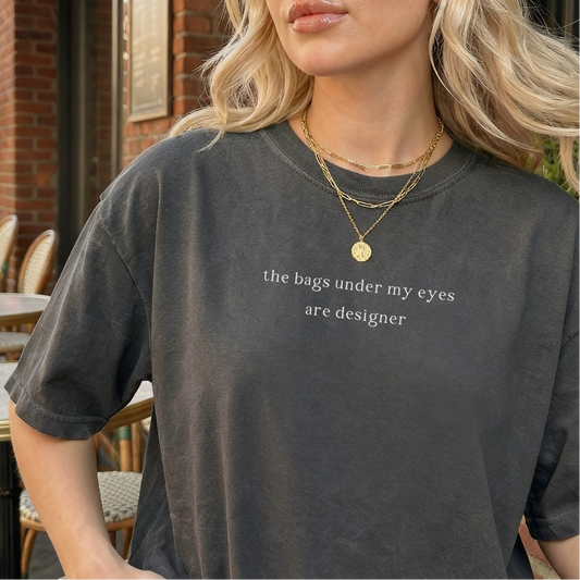 Woman wearing a pepper grey 'the bags under my eyes are designer' oversized t-shirt. She is accessorized with a layered gold necklace. Aesthetic and casual style photography for the Barely Sane clothing collection.