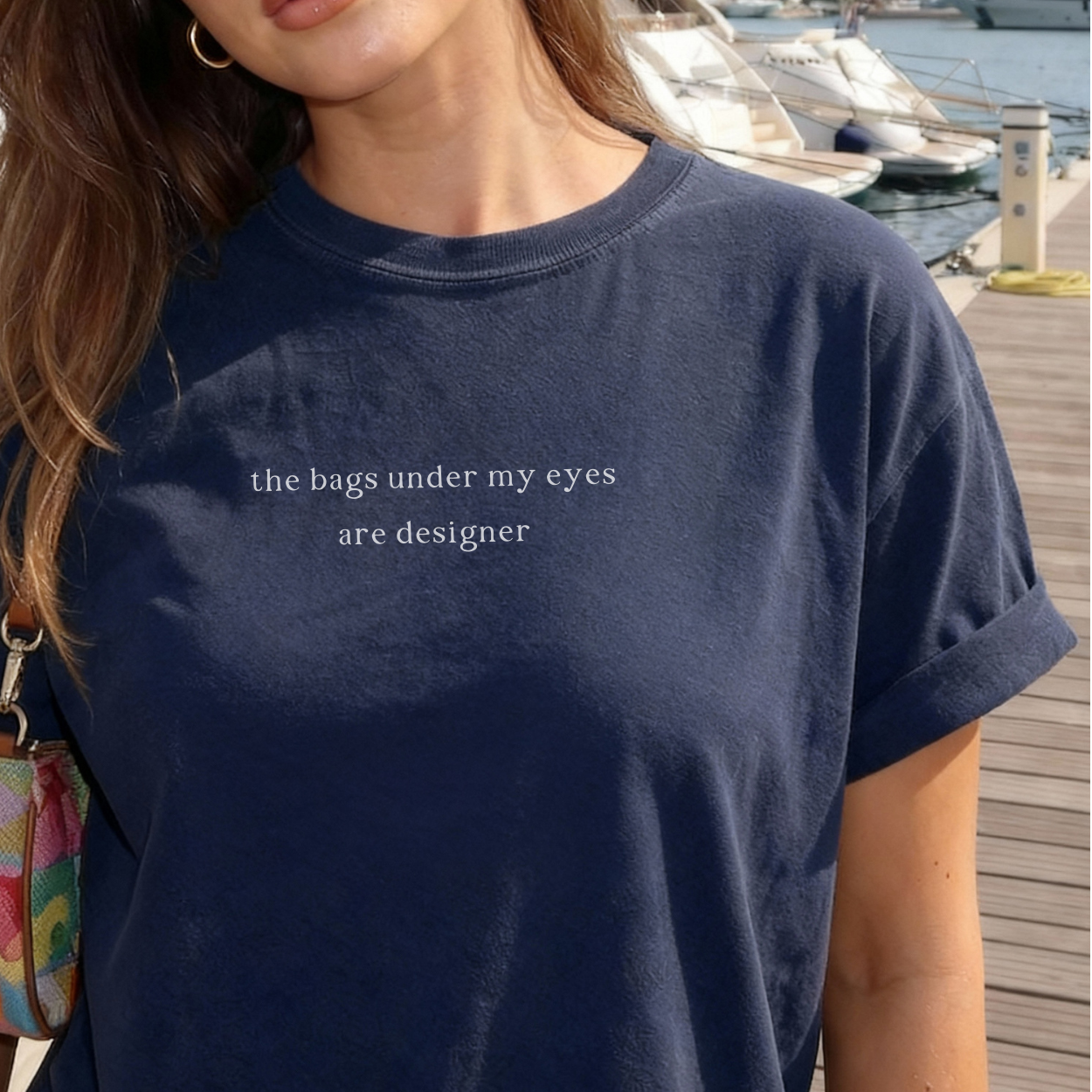 A close-up of a woman wearing a navy blue 'the bags under my eyes are designer' t-shirt. She is wearing gold hoop earrings. Relatable fashion, lifestyle photography for the Barely Sane clothing collection.
