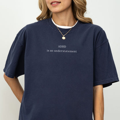 Oversized washed navy blue t-shirt with minimalist white text that reads 'ADHD is an understatement'. Model wears a simple gold chain necklace, showcasing the relaxed, vintage streetwear fit against a clean white background.