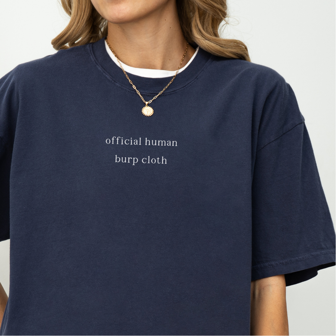 A close-up of a woman wearing a navy blue oversized t-shirt with the white text 'official human burp cloth' printed on the chest. She is wearing a gold coin necklace. Minimalist studio product photography for the Barely Sane collection.