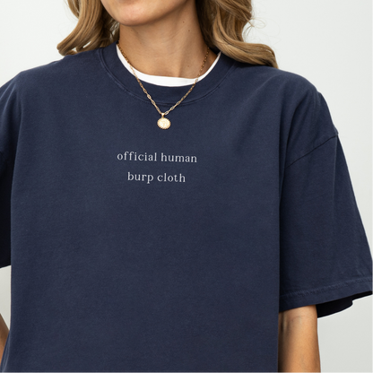 A close-up of a woman wearing a navy blue oversized t-shirt with the white text 'official human burp cloth' printed on the chest. She is wearing a gold coin necklace. Minimalist studio product photography for the Barely Sane collection.