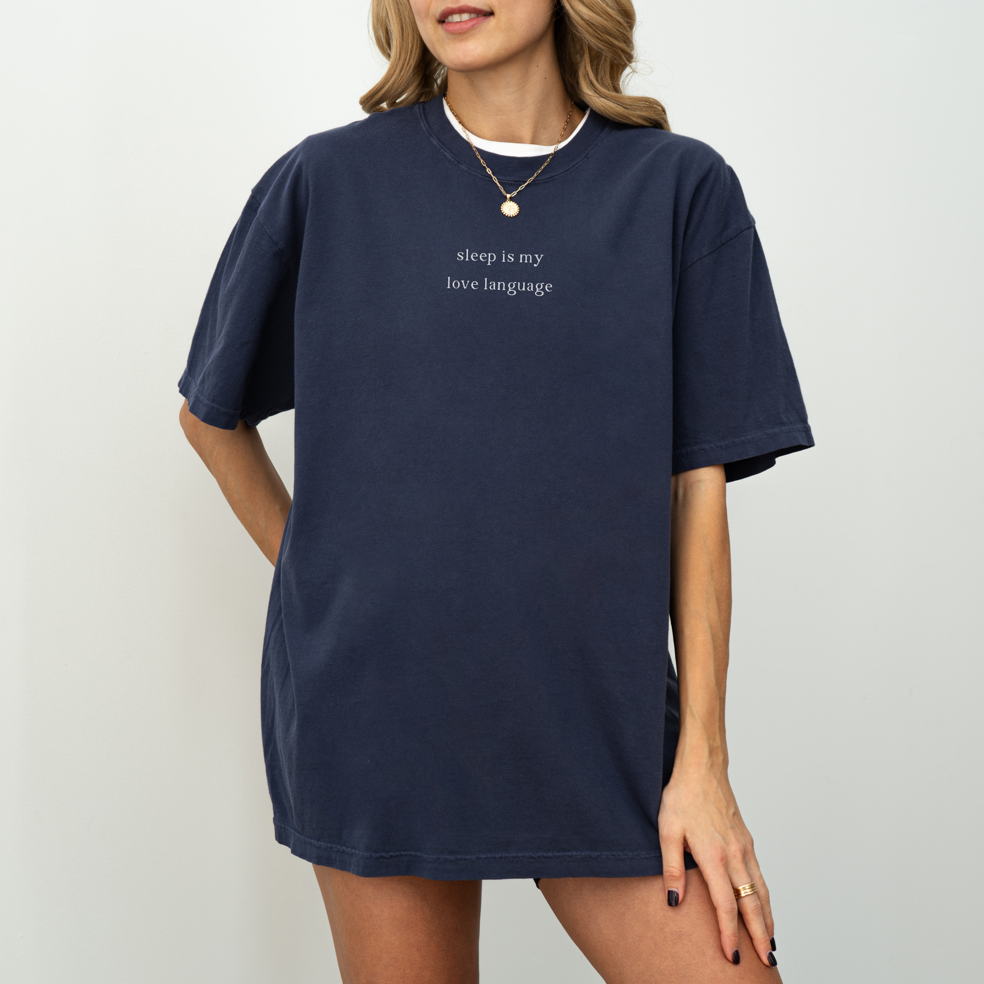 A woman wearing a navy blue 'sleep is my love language' oversized t-shirt, accessorized with a gold necklace and a red string bracelet. Minimalist studio product photography for the Barely Sane clothing collection.