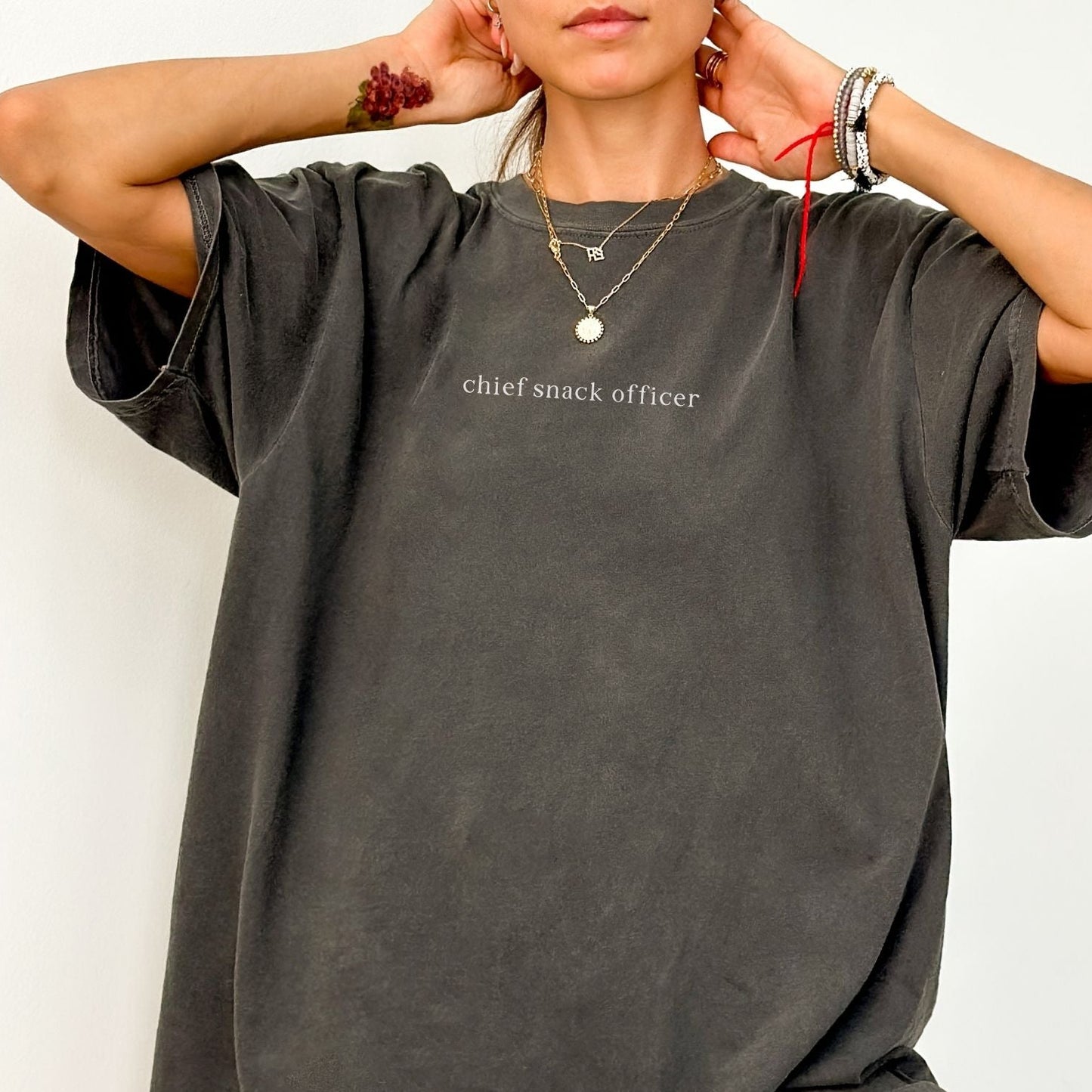 A close-up of a woman wearing a dark grey 'Chief Snack Officer' t-shirt. She is accessorized with layered gold necklaces and bracelets. High-quality, minimalist studio product photography for Barely Sane.