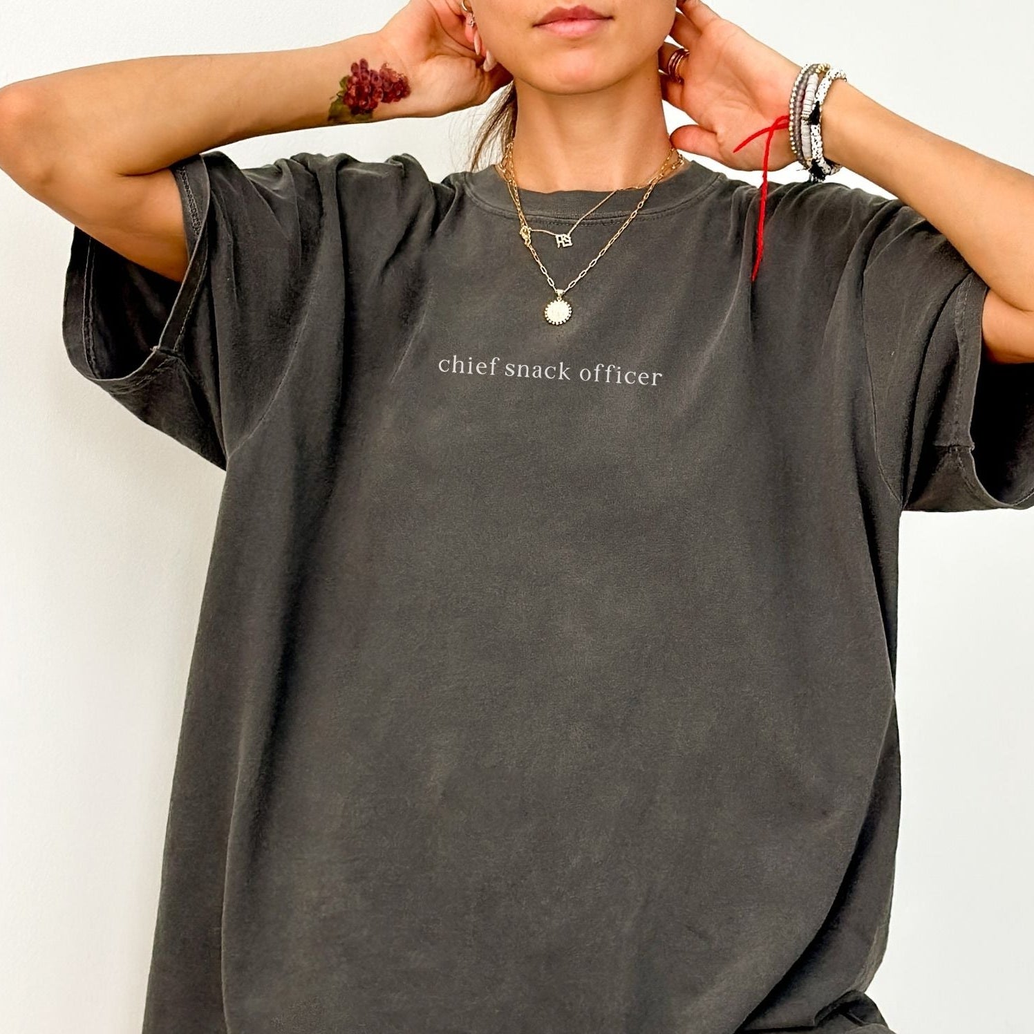 A close-up of a woman wearing a dark grey 'Chief Snack Officer' t-shirt. She is accessorized with layered gold necklaces and bracelets. High-quality, minimalist studio product photography for Barely Sane.
