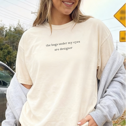 A woman smiling and wearing an ivory 'the bags under my eyes are designer' oversized t-shirt with a grey hoodie draped over her shoulders. Casual, aesthetic street style photography for the Barely Sane collection.
