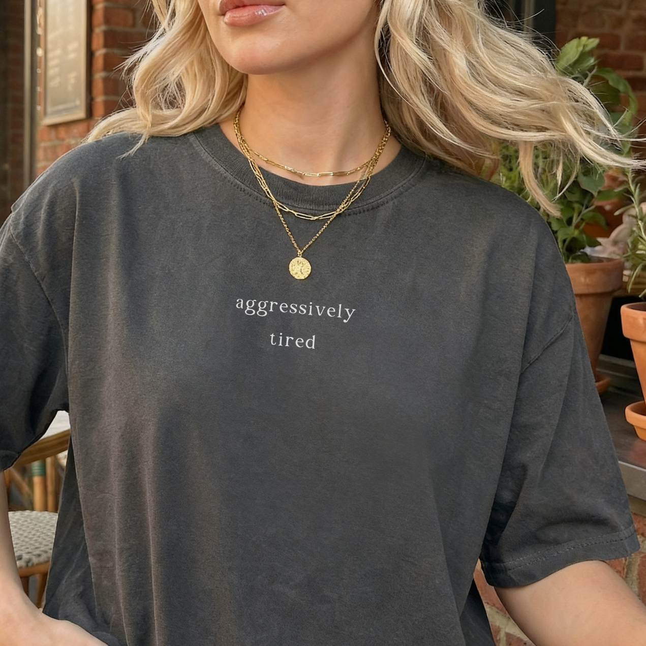 A woman wearing a pepper grey 'aggressively tired' oversized t-shirt. She is accessorized with layered gold necklaces. Close-up, aesthetic product photography for the Barely Sane clothing collection.