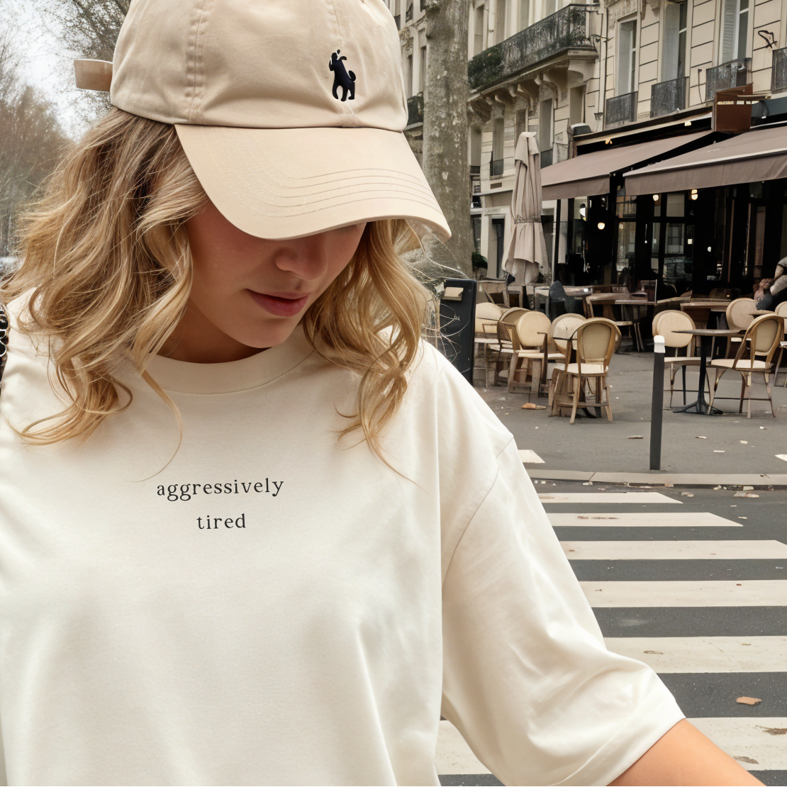 A woman wearing a tan baseball cap and an ivory 'aggressively tired' oversized t-shirt. She is looking down in a chic outdoor cafe setting with a Parisian street vibe. Aesthetic lifestyle photography for the Barely Sane clothing collection.