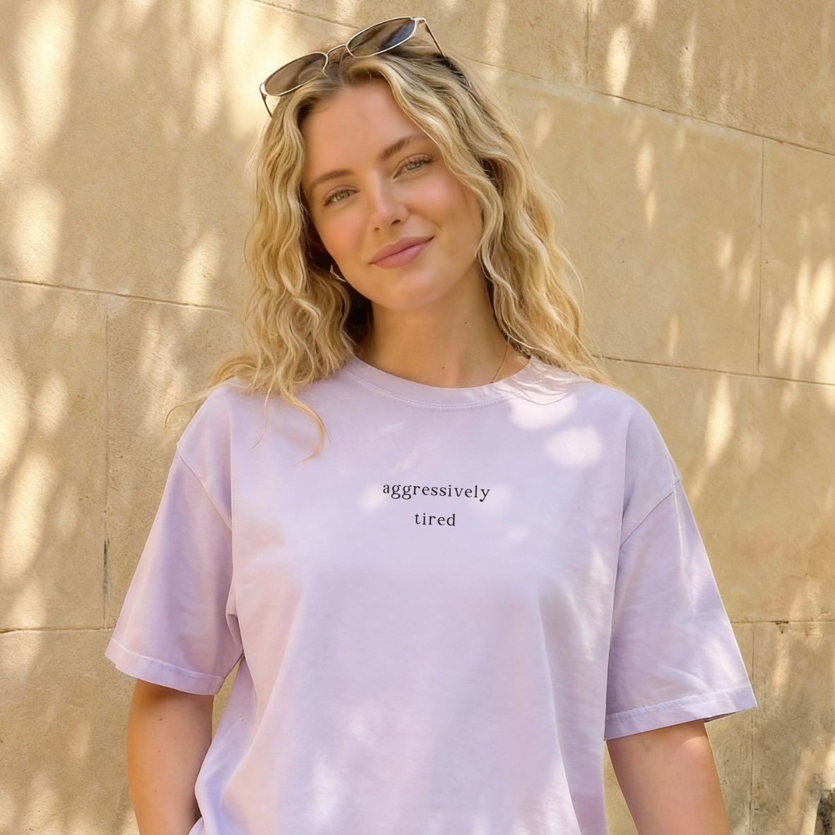 A woman wearing a light purple 'aggressively tired' t-shirt with sunglasses perched on her head. The background is a stone wall. Casual, aesthetic style photography for the Barely Sane clothing collection.