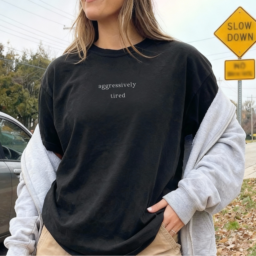 A woman wearing a black 'aggressively tired' oversized t-shirt with a grey hoodie draped over her shoulders and khaki pants. Casual, relatable lifestyle photography for the Barely Sane collection.