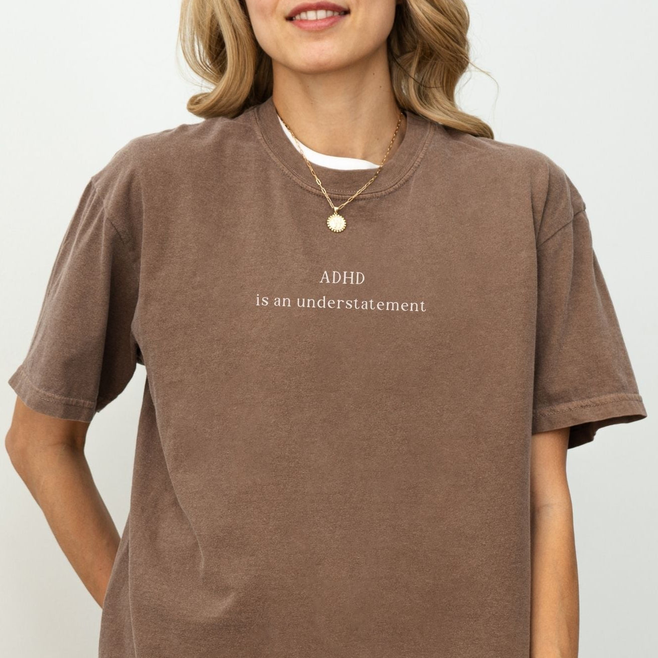 Oversized washed cocoa brown t-shirt with minimalist white text that reads 'ADHD is an understatement'. Blonde model wears a simple gold chain necklace, highlighting the vintage, aesthetic streetwear fit against a clean white background.