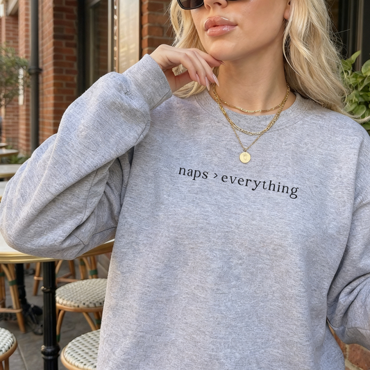 A woman wearing a sport grey sweatshirt with the black text 'naps > everything' printed on the chest. She is accessorized with layered gold necklaces in an outdoor cafe setting. Aesthetic lifestyle photography for the Barely Sane collection.
