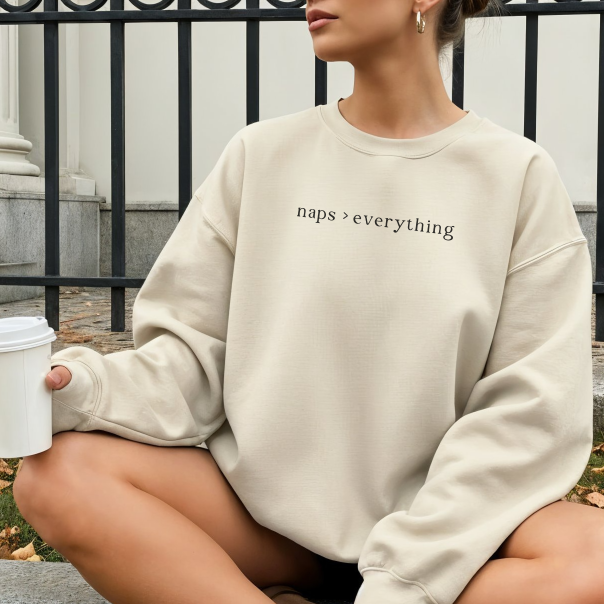 A woman sitting outdoors wearing a sand-colored oversized sweatshirt with the black text 'naps > everything' printed on the chest. She is holding a white coffee cup. Casual lifestyle photography for the Barely Sane collection.