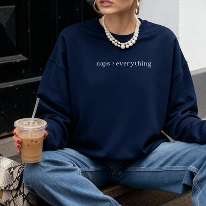 A woman wearing a navy blue sweatshirt with white text 'naps > everything' printed on the chest. She is accessorized with layered white bead necklaces and wearing blue jeans, holding a cup of iced coffee. Casual street style photography for the Barely Sane collection.