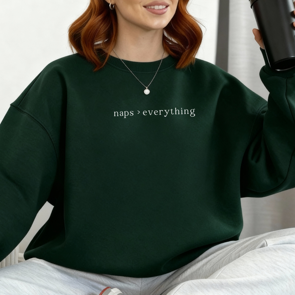 A woman with auburn hair wearing a forest green oversized sweatshirt with the white text 'naps > everything' printed on the chest. Casual lifestyle photography for the Barely Sane clothing collection.