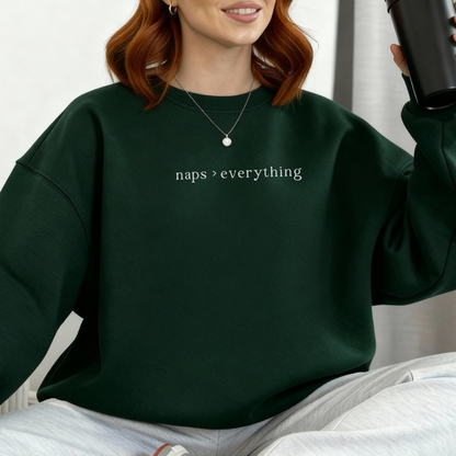 A woman with auburn hair wearing a forest green oversized sweatshirt with the white text 'naps > everything' printed on the chest. Casual lifestyle photography for the Barely Sane clothing collection.