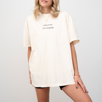 A woman wearing an ivory 'sleep is my love language' oversized t-shirt, accessorized with a gold necklace and a red string bracelet. Minimalist studio product photography for the Barely Sane clothing collection.
