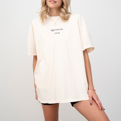 A woman wearing an ivory 'aggressively tired' oversized t-shirt. She is accessorized with a gold necklace and a gold ring. Minimalist studio product photography for the Barely Sane clothing collection.