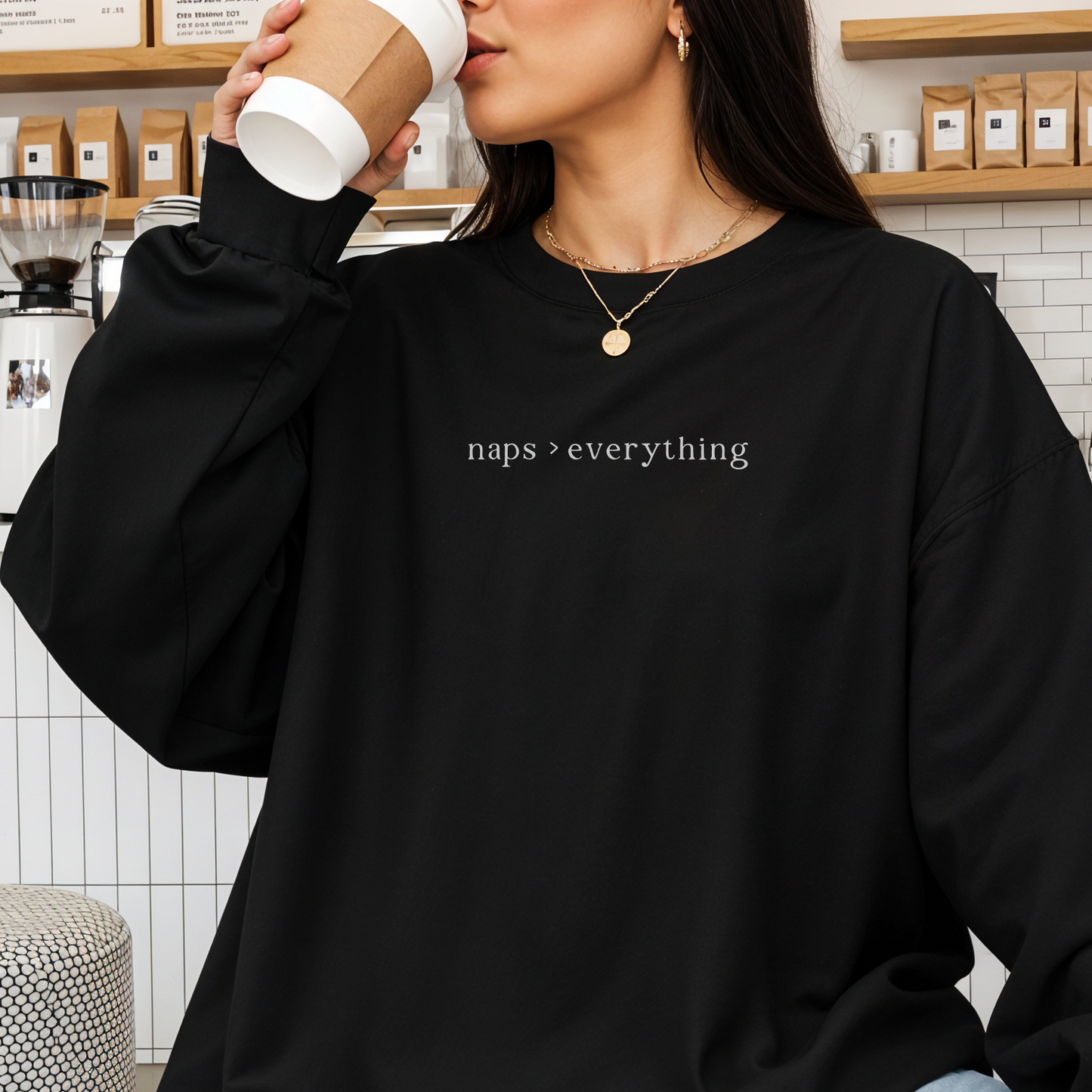 A close-up of a woman in a cafe wearing a black oversized sweatshirt with the white text 'naps > everything' printed on the chest. She is holding a coffee cup and accessorized with layered gold necklaces. Aesthetic lifestyle photography for the Barely Sane collection.