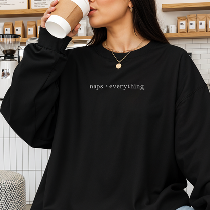 A close-up of a woman in a cafe wearing a black oversized sweatshirt with the white text 'naps > everything' printed on the chest. She is holding a coffee cup and accessorized with layered gold necklaces. Aesthetic lifestyle photography for the Barely Sane collection.