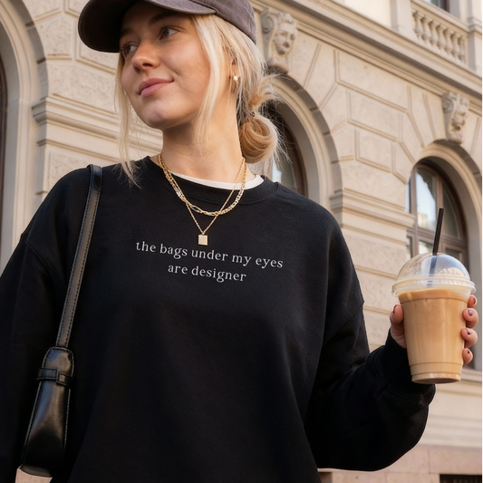 A woman wearing a black oversized sweatshirt with white text on the chest that reads 'the bags under my eyes are designer'. She is carrying an iced coffee and a black shoulder bag, and wearing a grey baseball cap and layered gold necklaces. Outdoors, classic architecture background. Casual lifestyle photography for Barely Sane.