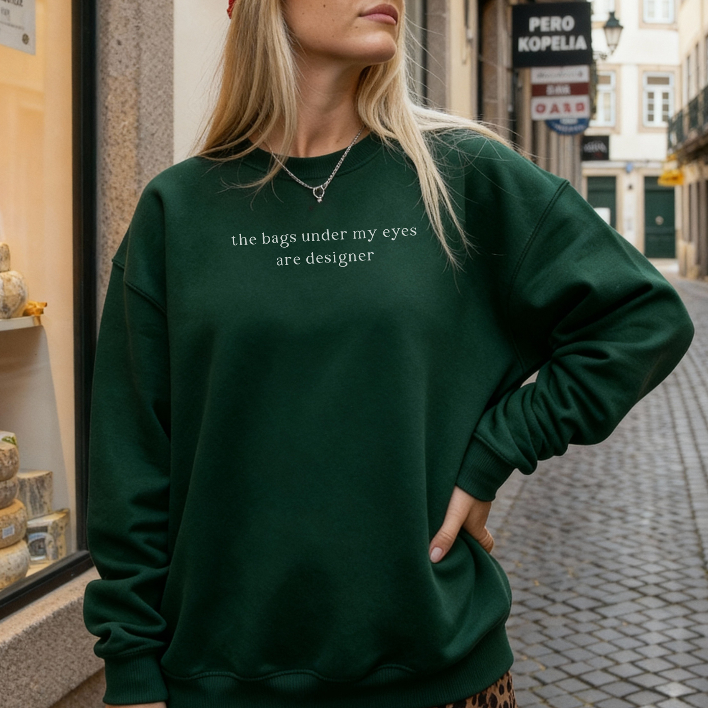 A woman with long blonde hair wearing a forest green sweatshirt with white text on the chest 'the bags under my eyes are designer'. Casual lifestyle street style photography in a European city for Barely Sane clothing.