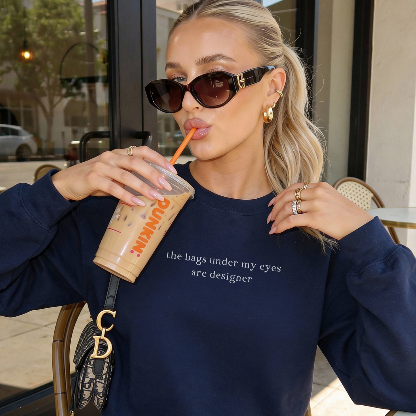 A blonde woman wearing a navy blue oversized sweatshirt with white text 'the bags under my eyes are designer'. She is sipping an iced coffee, wearing dark sunglasses and gold jewelry, with a designer shoulder bag visible. Aesthetic lifestyle photography for Barely Sane.