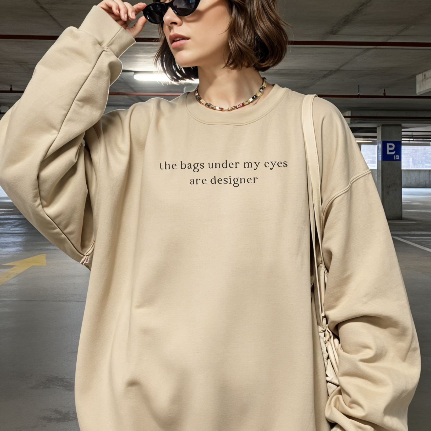 A woman wearing a sand-colored oversized sweatshirt with the black text 'the bags under my eyes are designer' printed on the chest. She is holding sunglasses and wearing a colorful beaded necklace in an underground parking garage. Aesthetic lifestyle photography for the Barely Sane collection.