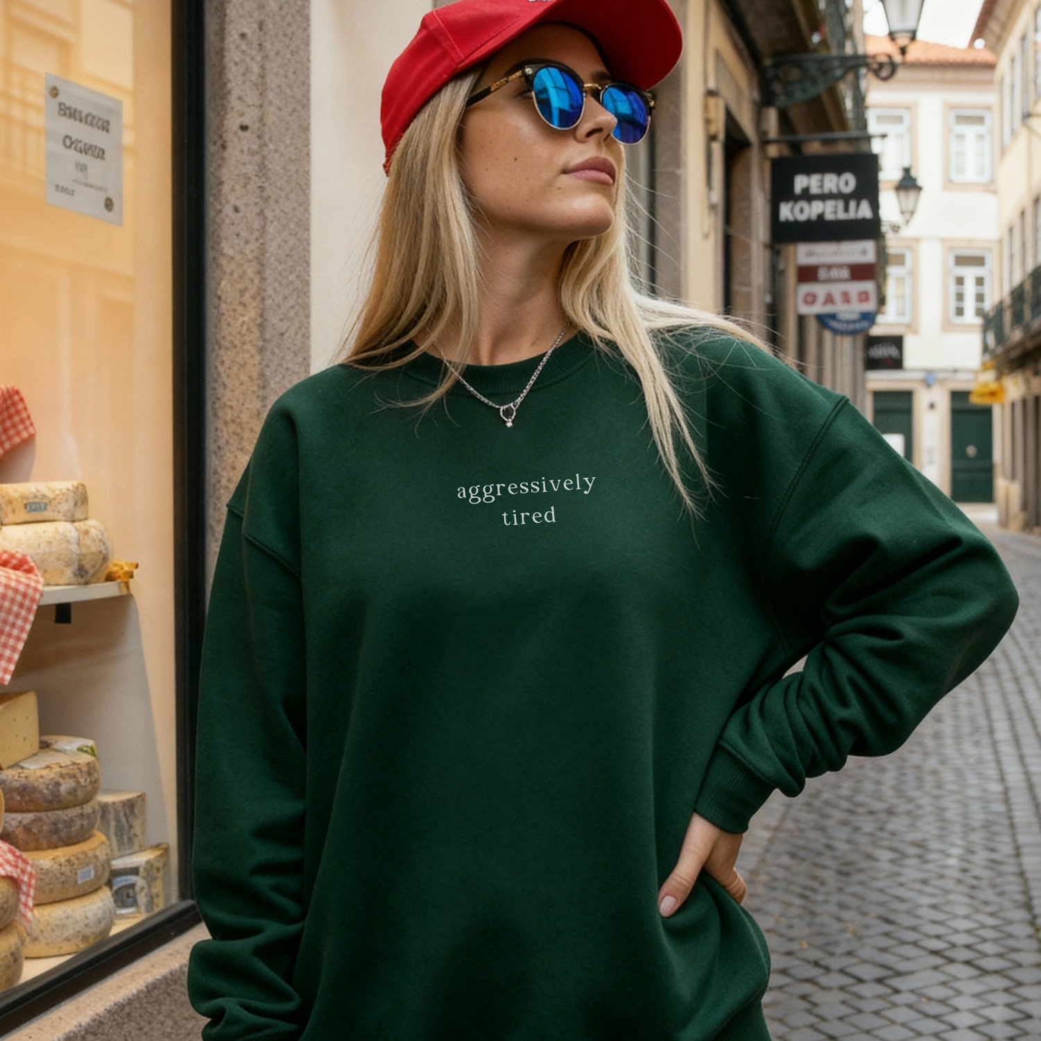 A woman wearing a forest green oversized sweatshirt with the white text 'aggressively tired' printed on the chest. She is accessorized with a red baseball cap and blue reflective sunglasses. Casual street style photography with a cheese shop background for Barely Sane.