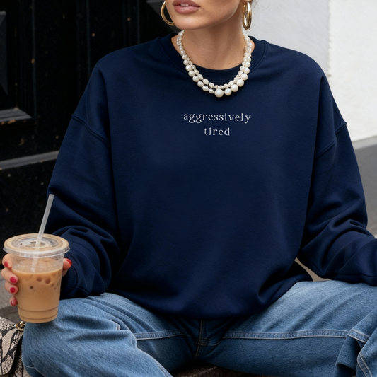A woman wearing a navy blue oversized sweatshirt with the white text 'aggressively tired' printed on the chest. She is accessorized with layered white bead necklaces and holding a cup of iced coffee. Casual lifestyle photography for the Barely Sane collection.