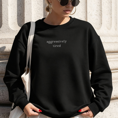 A woman wearing a black oversized sweatshirt with the white text 'aggressively tired' printed on the chest. She is also carrying a natural canvas tote bag and wearing black sunglasses and gold hoop earrings. Outdoors, architectural background. Casual lifestyle photography for Barely Sane.