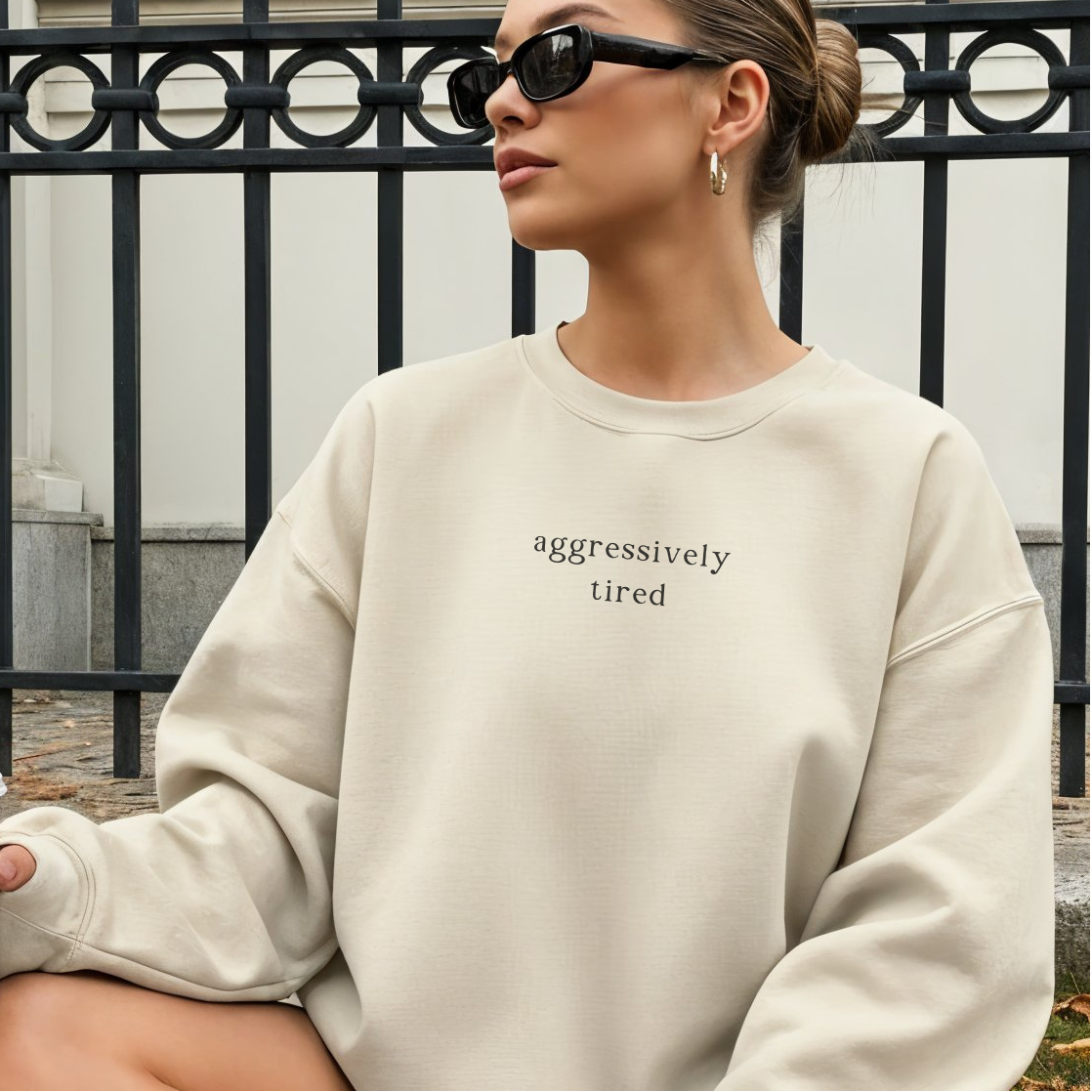 A woman wearing a sand-colored oversized sweatshirt with the black text 'aggressively tired' printed on the chest. She is wearing black sunglasses and gold hoop earrings, posed in an outdoor setting. Aesthetic lifestyle photography for the Barely Sane collection.