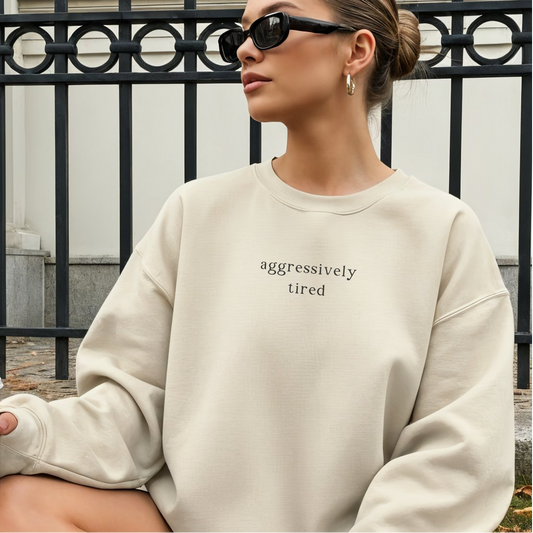 A woman wearing a sand-colored oversized sweatshirt with the black text 'aggressively tired' printed on the chest. She is wearing black sunglasses and gold hoop earrings, posed in an outdoor setting. Aesthetic lifestyle photography for the Barely Sane collection.
