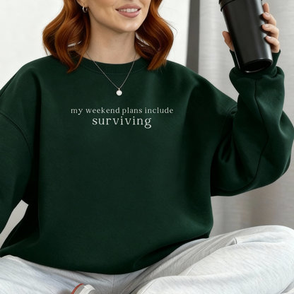 a woman wearing a forest green sweatshirt with text 'my weekend plans include surviving' styled with light grey seatpants and a gold necklace