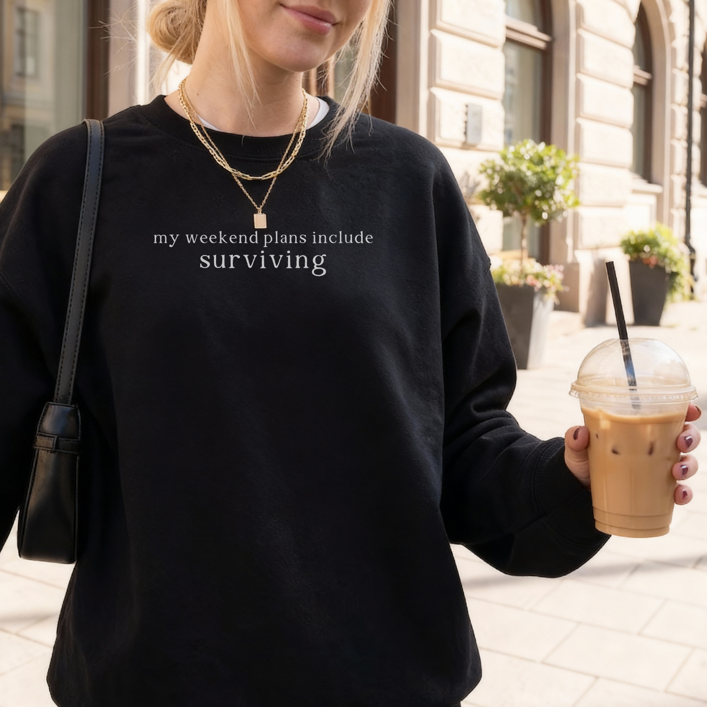 a woman wearing a black sweatshirt with text 'my weekend plans include surviving' styled with gold necklaces, holding a cup of cold coffee