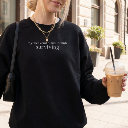 a woman wearing a black sweatshirt with text 'my weekend plans include surviving' styled with gold necklaces, holding a cup of cold coffee