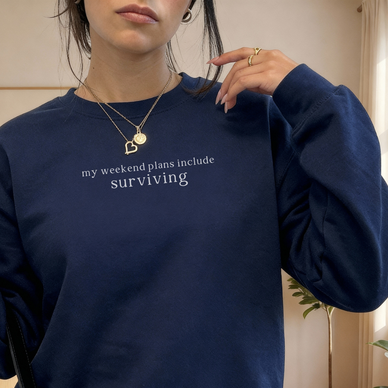 a woman wearing a navy blue sweatshirt with text 'my weekend plans include surviving' styled with gold necklaces and rings