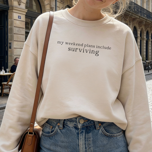 a woman wearing a beige sweatshirt with text 'my weekend plans include surviving' in an urban setting.